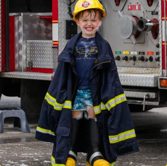 Smiling kid in firefighter gear by a real fire truck